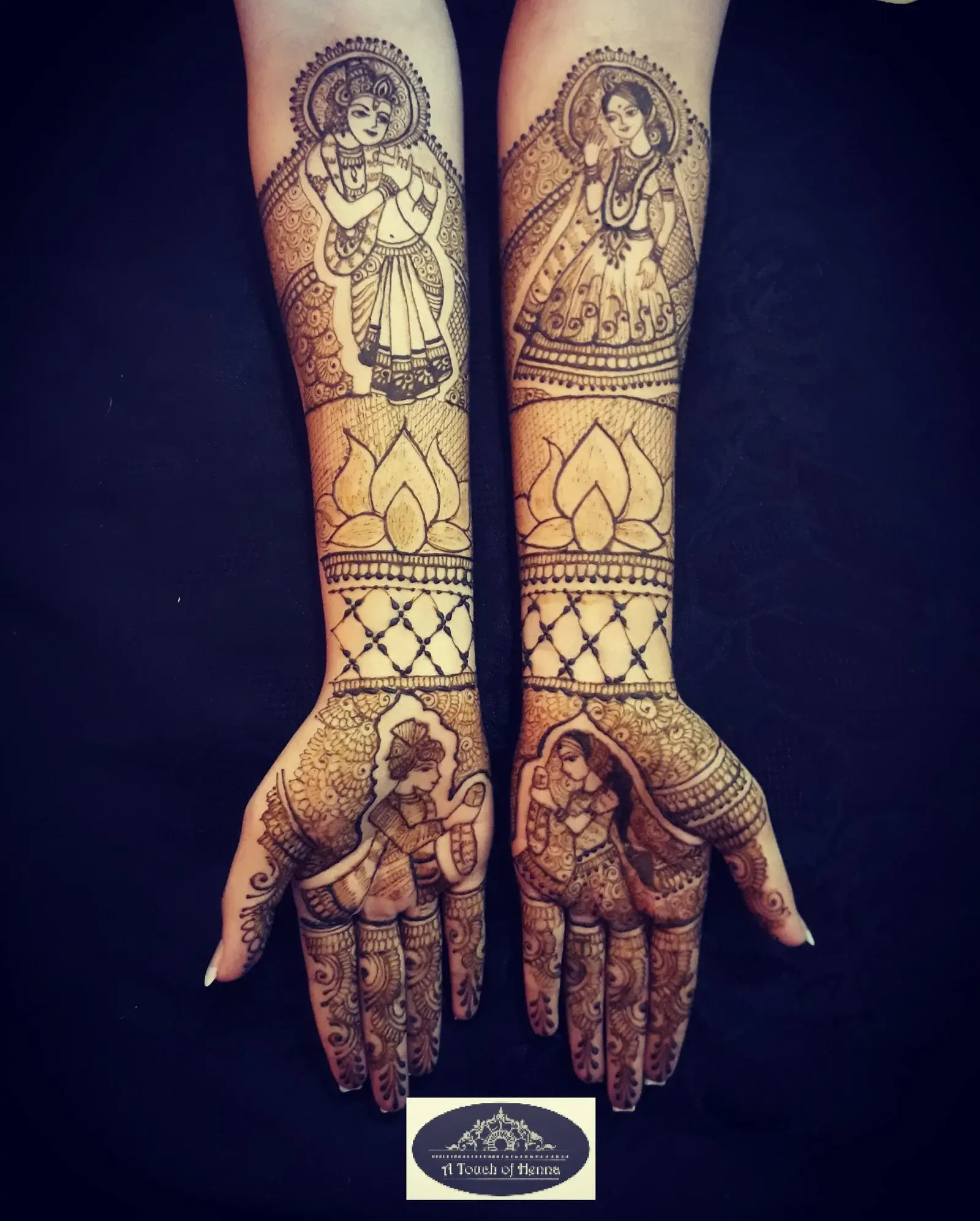 Elaborate full-arm bridal mehndi with deity figures, lotus flowers and lattice patterns on both arms against dark background